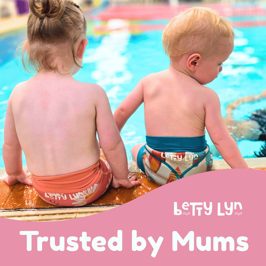 Two young children sitting at the edge of an indoor swimming pool, viewed from behind. The child on the left wears a peach-pink one-piece swimsuit, and the child on the right wears a multicolor swim brief with a teal waistband.